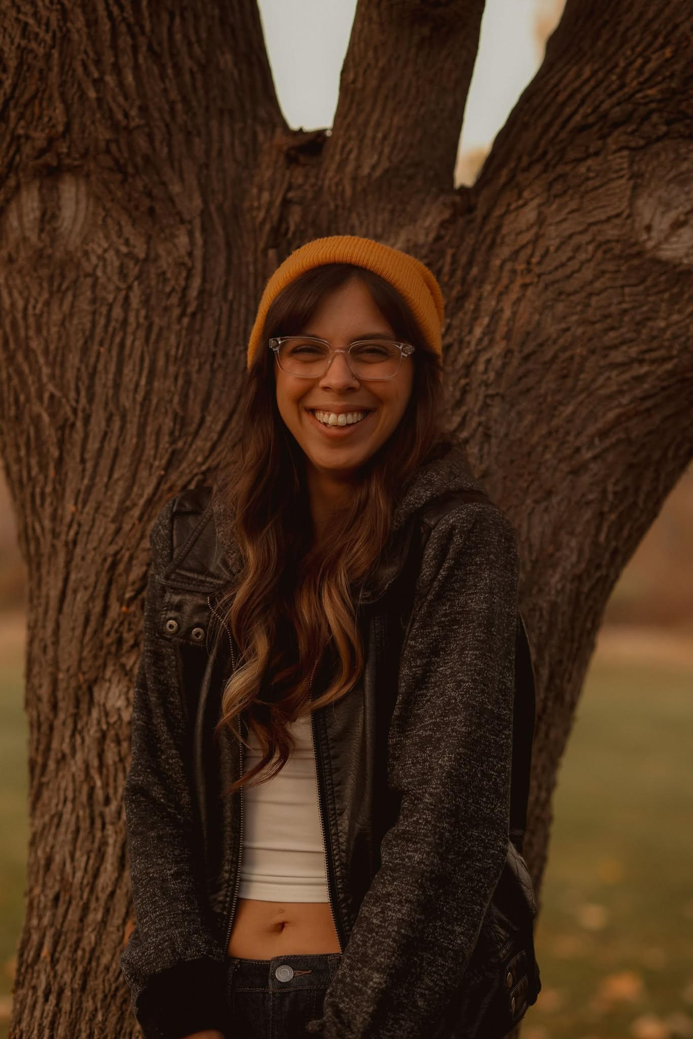 An image of Kayla Henshaw, owner of the Fig Leaf Cafe. With long dark, curly hair, glasses, a yellow beanie, a white crop top and jeans. Posed in front of a tree.